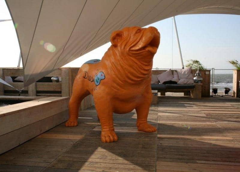 Large bulldog figure 1 Pomarańczowa statua psa na drewnianym tarasie pod białym zadaszeniem.