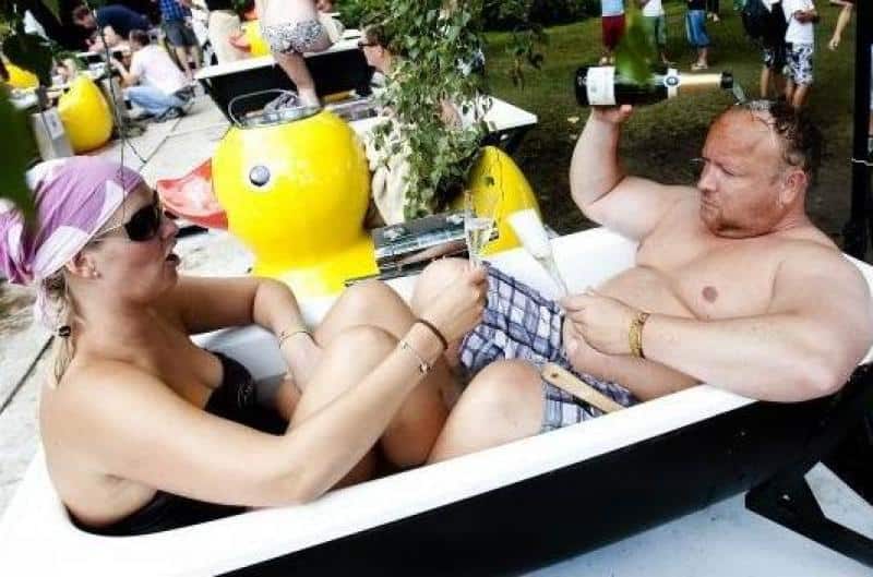 oryg_064490685_1129949550544944_1465385417682452480_o A man and woman, wearing swimsuits, relaxing in a filled bathtub outdoors. The man is holding a bottle of champagne while the woman holds a glass. Several other people and bathtubs can be seen in the background.