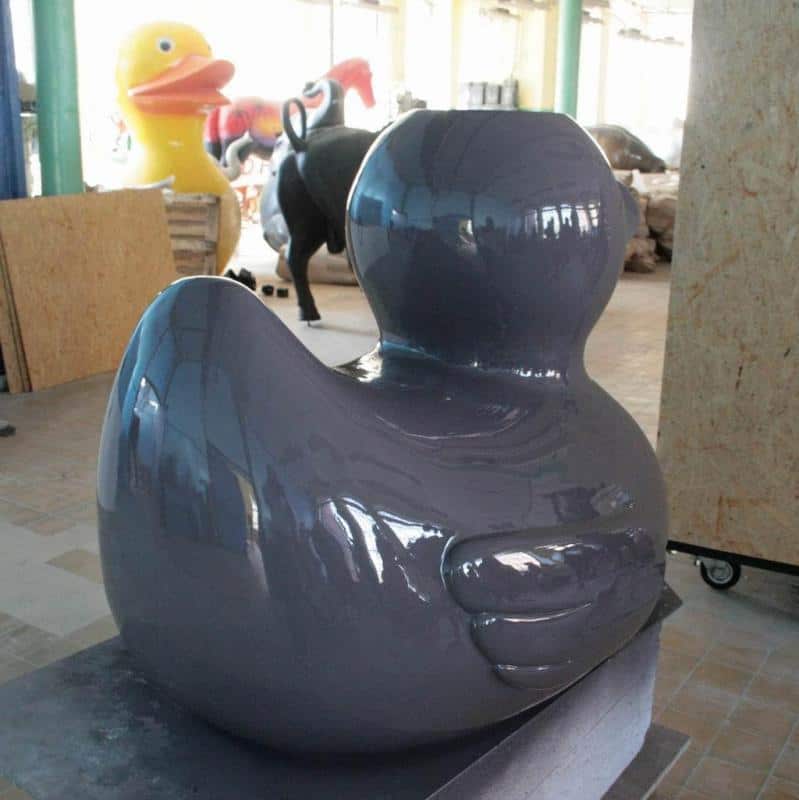 Festival Attractions 3 Duża, szara rzeźba kaczki w warsztacie z większymi rzeźbami w tle, w tym żółtą kaczką.