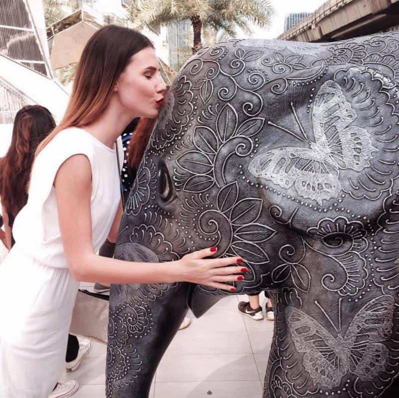 oryg_064490685_1129949550544944_1465385417682452480_o Woman in white dress kissing a decorative elephant statue adorned with intricate patterns and butterfly designs.