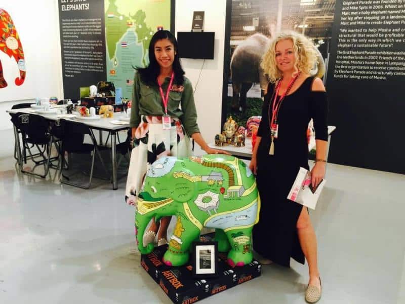 Elephant Parade 2 Dwie kobiety stojące obok kolorowej figurki słonia, trzymające broszury na tle wystawy.