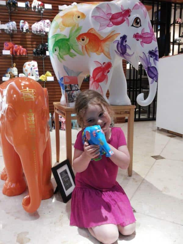 Elephant Parade 3 Dziewczynka w różowej sukience, trzymająca kolorową figurkę słonia, siedząca przy większych, pomalowanych słoniach dekoracyjnych.