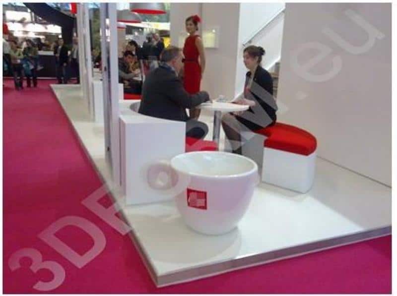 oryg_064490685_1129949550544944_1465385417682452480_o People sitting at a booth in a trade show, with a pink carpet, white and red furniture, and a woman standing in the background.