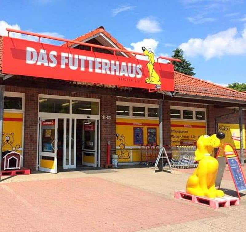 FUTTERHAUS Advertising Dog 2 Sklep Das Futterhaus z czerwonym szyldem nad wejściem, żółtą figurką psa i plakatami.