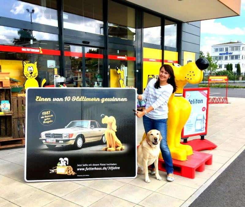 oryg_064490685_1129949550544944_1465385417682452480_o Woman and dog standing outside a store, holding a large sign about winning one of 10 old-timers, with a car and cartoon dog image.