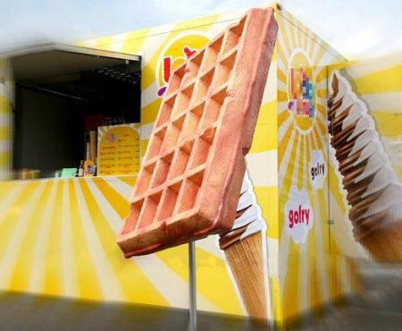 Large waffle on a stick in front of a yellow food truck with ice cream cone graphics.