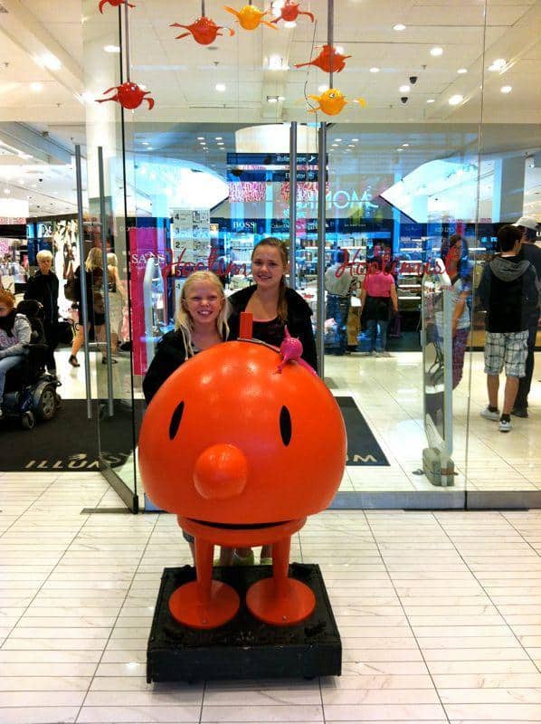 oryg_064490685_1129949550544944_1465385417682452480_o Two people posing with a large, orange, round character statue inside a shopping mall.