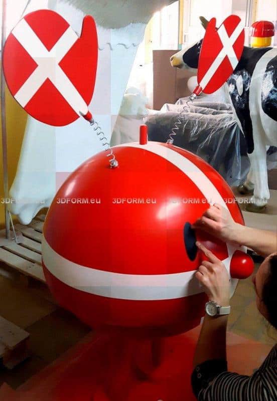 Hoptimist Advertising Figure 1 Czerwona, okrągła figura z uszami na sprężynach i białymi paskami, z rękami przymocowującymi oko. W tle plastikowa krowa.