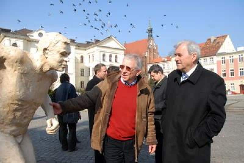 IAAF World Championships Promotion 1 Dwóch mężczyzn rozmawiających obok rzeźby na rynku miejskim, w tle kamienice i wieża zegarowa.