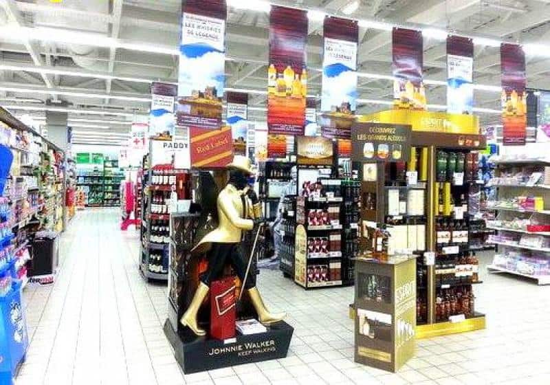 Supermarket aisle with extensive liquor displays, including prominent Johnnie Walker and Ricard stands, surrounded by shelves of alcoholic beverages.