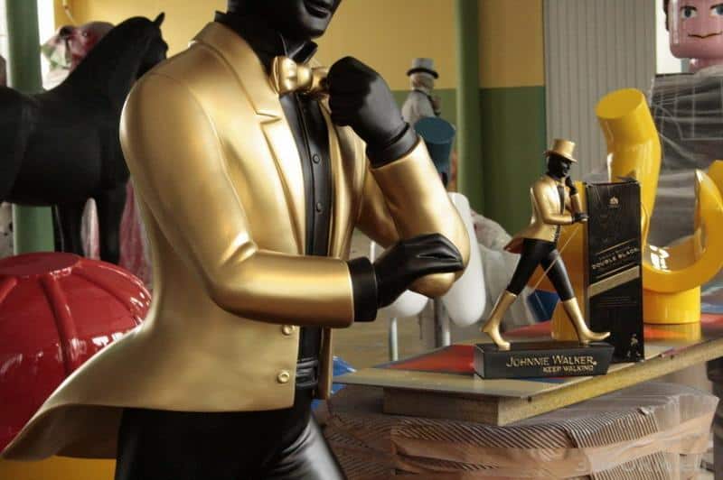 Złota figurka Johni Walker w złotym garniturze, obok mniejsza statuetka i pudełko.