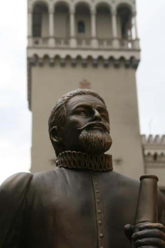 oryg_064490685_1129949550544944_1465385417682452480_o Statue of a historical figure with a beard and ruffed collar holding a scroll, with a tower in the background.
