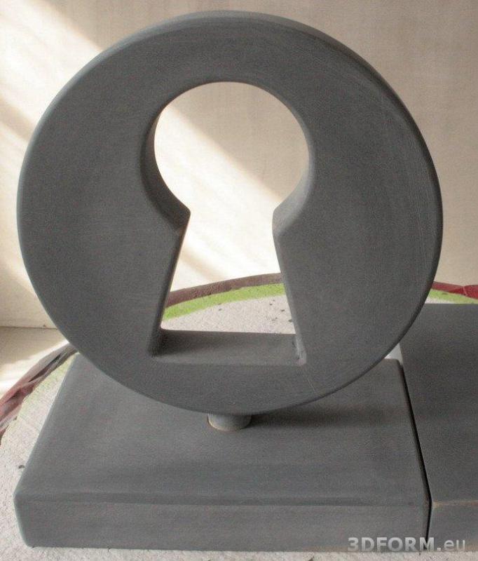 Keyhole / Statuette 2 Szara rzeźba w kształcie zamka do klucza, umieszczona na prostokątnej podstawie.