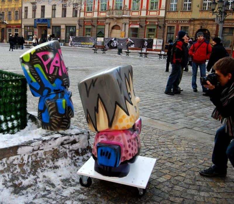 Large art sculptures resembling stylized cartoon characters on a cobblestone city square, with a person taking a photo and several people walking in the background.