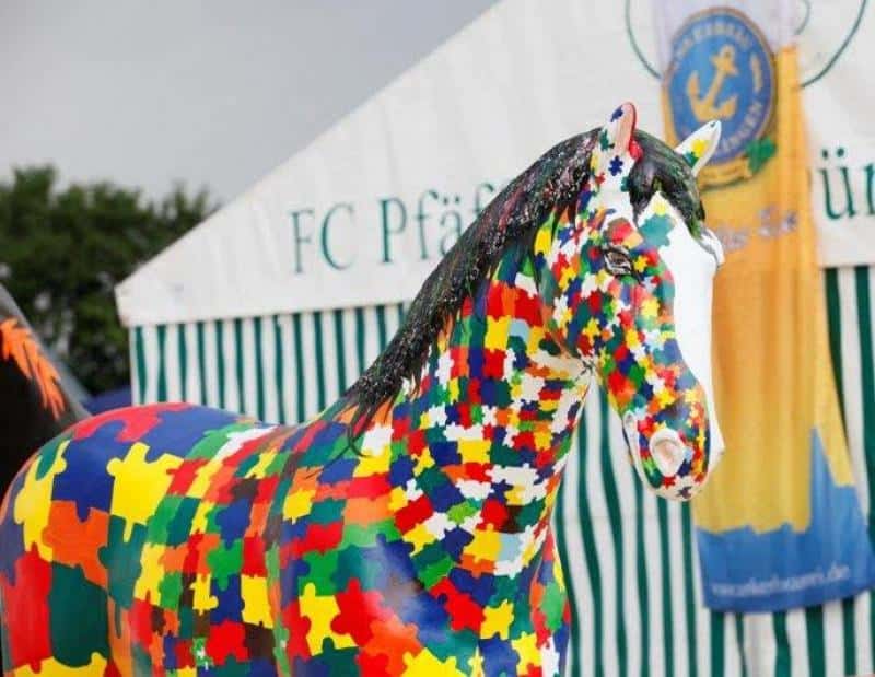 oryg_064490685_1129949550544944_1465385417682452480_o Colorful horse statue painted with puzzle piece pattern, standing in front of a striped tent.