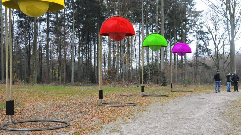 oryg_064490685_1129949550544944_1465385417682452480_o Colorful dome-shaped outdoor installations in yellow, red, green, and purple suspended on poles in a forest area with two people observing.