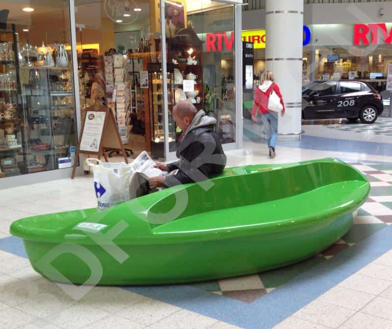 oryg_064490685_1129949550544944_1465385417682452480_o A man sitting on a bright green bench shaped like a boat in a shopping mall, reading a newspaper.