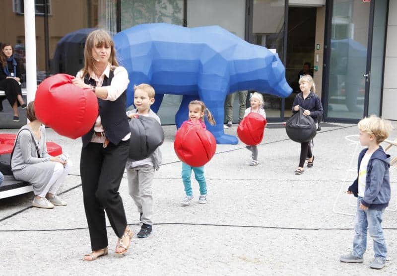 oryg_064490685_1129949550544944_1465385417682452480_o Children and an adult carrying large, round, red and black cushions in an outdoor setting with a blue polygonal bear sculpture in the background.