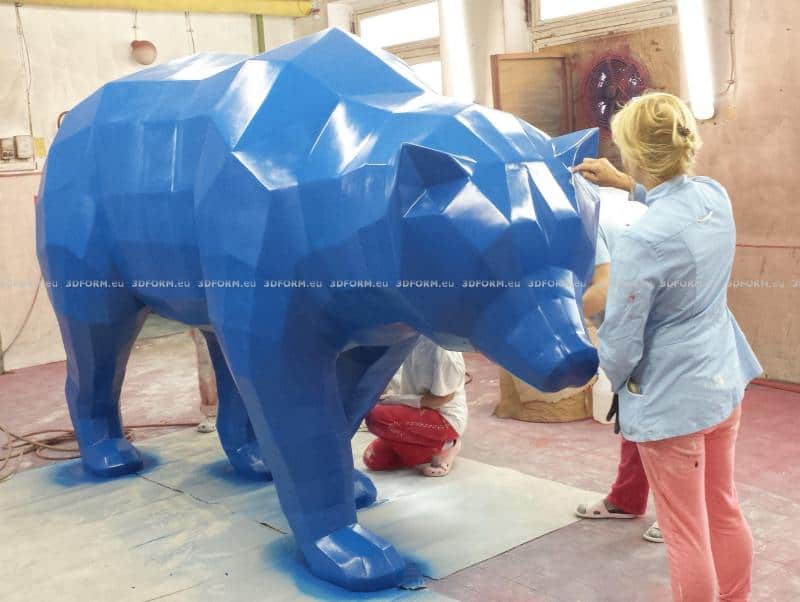 Low-poly geometric bear 1 Kobieta maluje dużą, niebieską, geometryczną rzeźbę niedźwiedzia w pracowni artystycznej.