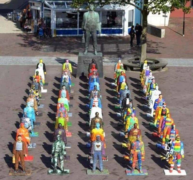 Kolorowe figury ludzi ustawione w szeregu na placu przed dużym pomnikiem mężczyzny.