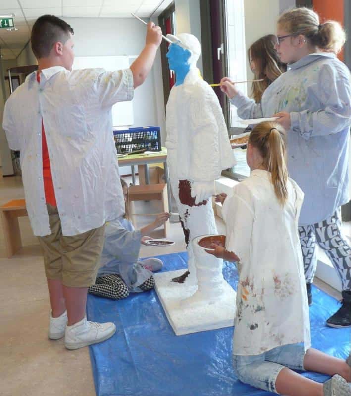 Dzieci malują stojącą rzeźbę człowieka w szkolnej sali.