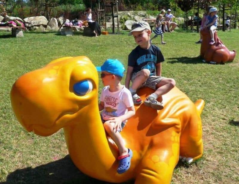 oryg_064490685_1129949550544944_1465385417682452480_o Two young children riding a large, orange dinosaur-shaped playground structure in a park.
