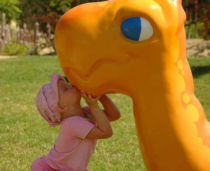 Dragons in the amusement park 1 Dziewczynka w różowym ubraniu całuje dużą, pomarańczową figurkę dinozaura na placu zabaw.