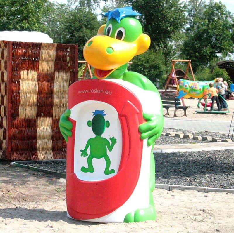 oryg_064490685_1129949550544944_1465385417682452480_o A colorful cartoonish trash can shaped like a green dinosaur holding a red bin, with a playground in the background.