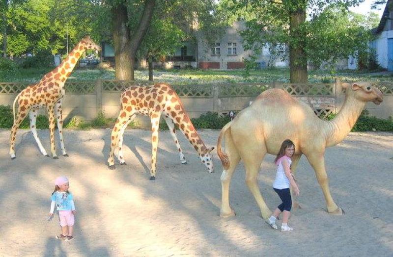 Figures in a theme park 4 Dzieci bawiące się obok figur żyraf i wielbłąda w parku.