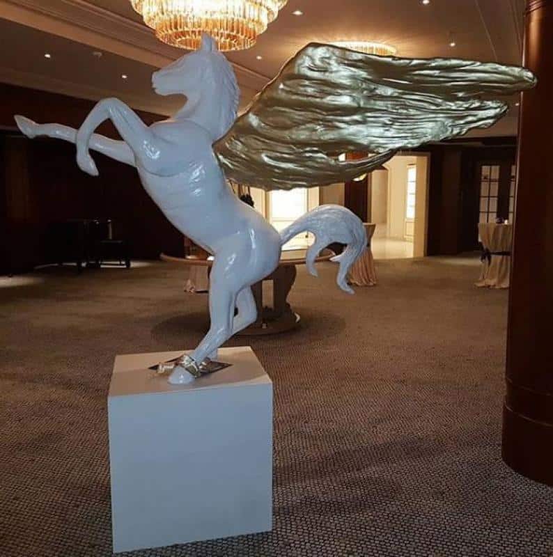 oryg_064490685_1129949550544944_1465385417682452480_o Statue of a white horse with golden wings rearing up, placed on a pedestal in an elegant room with chandeliers and a carpeted floor.