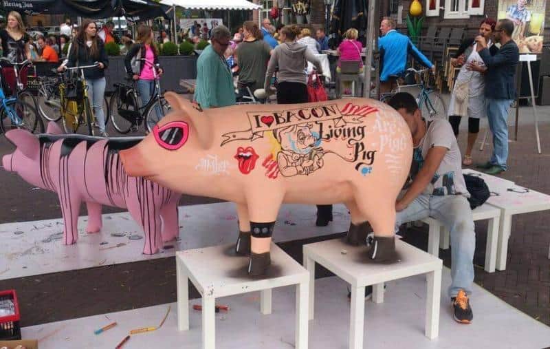 oryg_064490685_1129949550544944_1465385417682452480_o People in an outdoor plaza painting cartoonish pig sculptures. One pig sculpture is painted with a tattoo-style design featuring the words