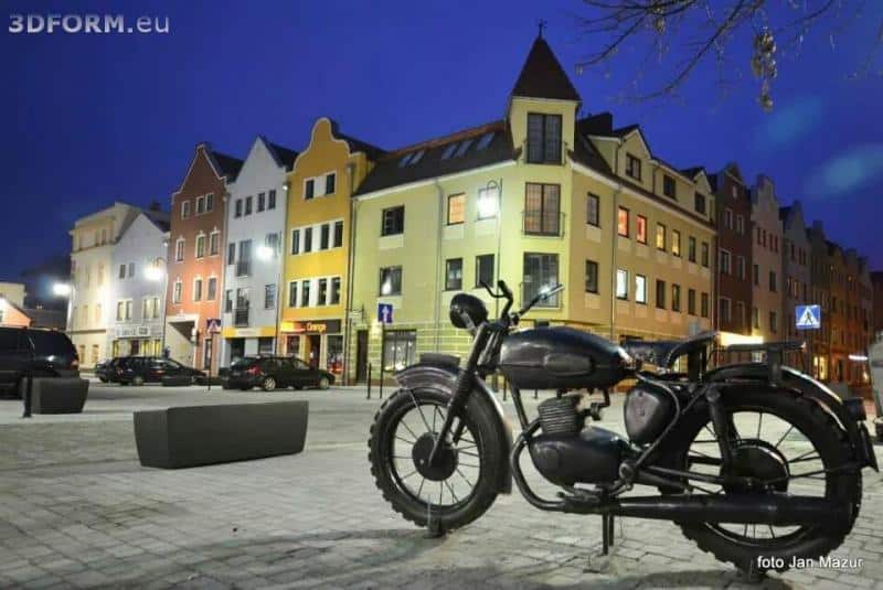 oryg_064490685_1129949550544944_1465385417682452480_o Vintage motorcycle parked in front of colorful buildings at dusk in a city square.