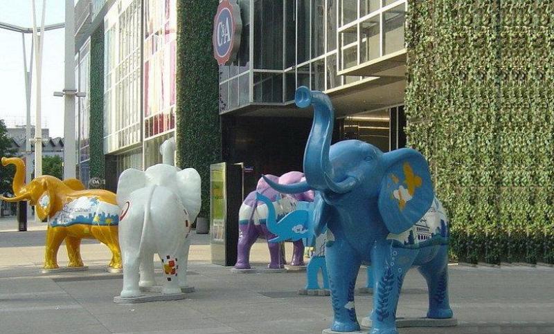 Colorful elephant statues displayed outdoors in front of a modern building with a green wall.