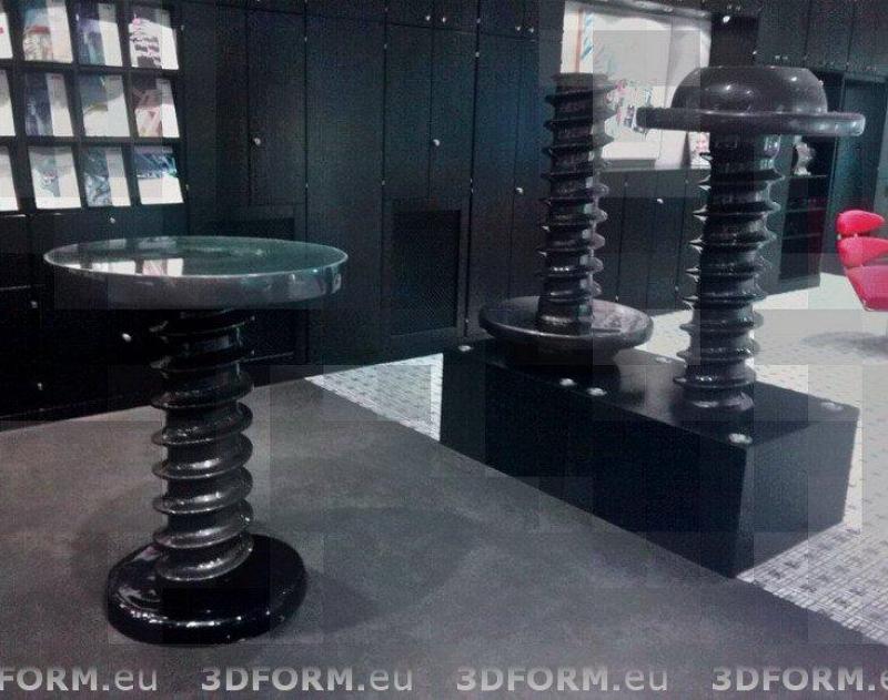 Three round tables in a modern setting, each with a thick, spiraled metal base and a black finish, with a glass top on one and dark tops on the other two. The background features black cabinets and a red chair.