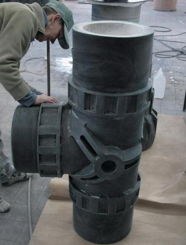 Tee - Pipe Fitting 3 Mężczyzna w warsztacie pochylający się nad dużym rzeźbionym modelem krzyżowego połączenia rur.