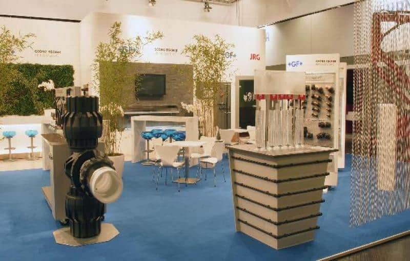 oryg_064490685_1129949550544944_1465385417682452480_o Trade show booth with industrial equipment displays, tall metal pipes, seating area with blue stools and white tables, plants, and a blue carpet.