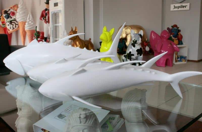 School of Tuna 3 Białe figurki rekinów na szklanym stole, w tle kolorowe figurki zwierząt i inne dekoracje.