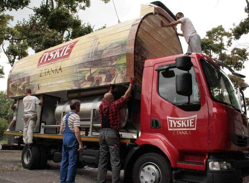 oryg_064490685_1129949550544944_1465385417682452480_o Workers securing a large wooden Tyskie beer barrel on a red truck.