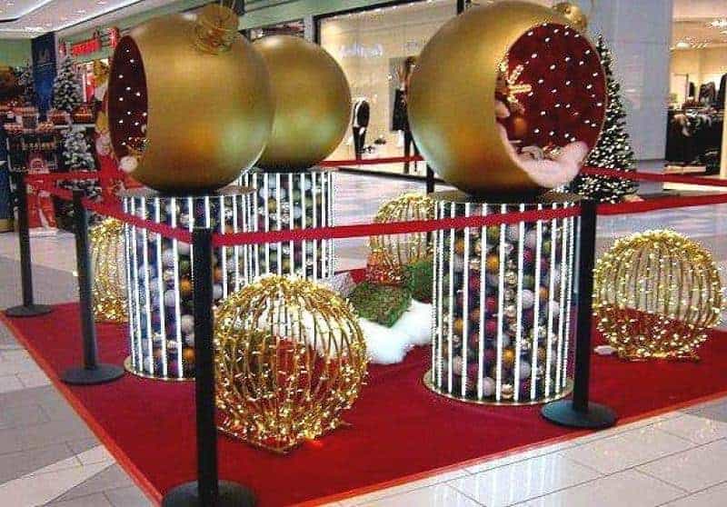 Illuminated Christmas baubles 4 Duże złote bombki świąteczne stojące na cylindrycznych podstawach, otoczone czerwoną taśmą w centrum handlowym.