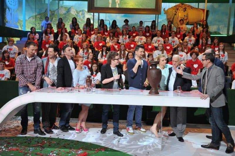 oryg_064490685_1129949550544944_1465385417682452480_o A group of people standing behind a long table on a colorful television set, with a large audience seated in the background.