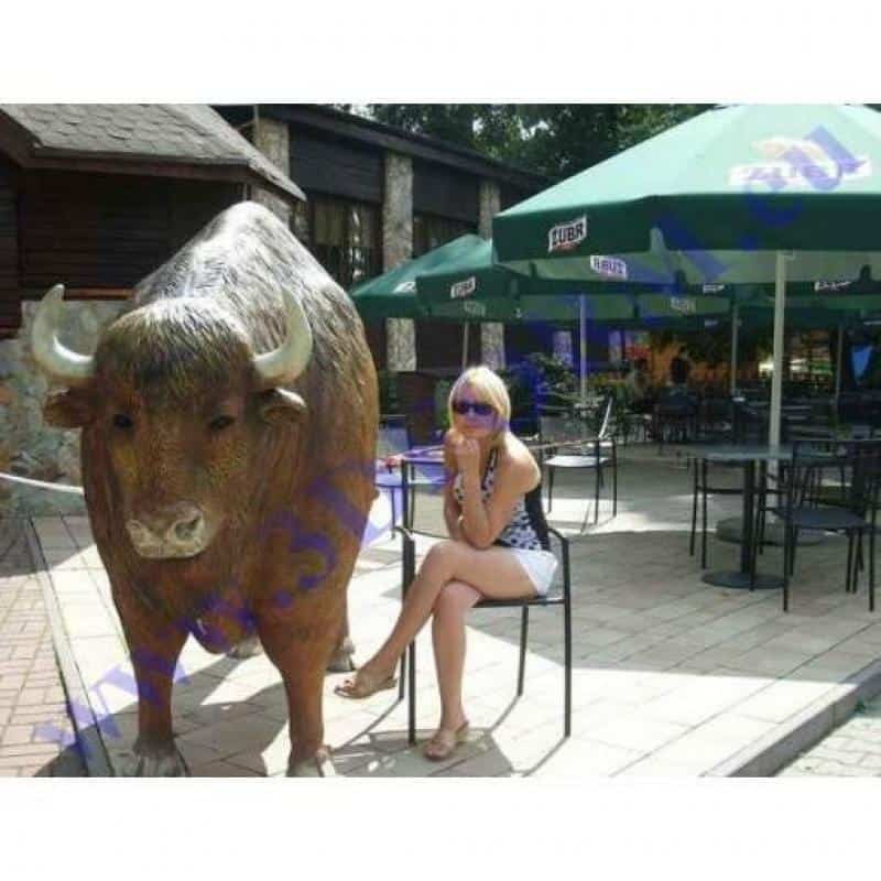 oryg_064490685_1129949550544944_1465385417682452480_o A person sitting on a chair next to a large bull statue outside a café with green umbrellas.