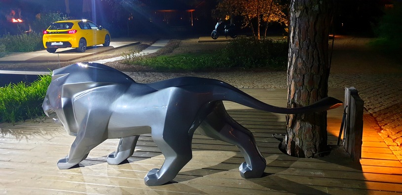 Peugeot lion fiberglass advertising sculpture 4 Figurka lwa w kolorze srebrnym na drewnianej platformie, w tle żółty samochód na oświetlonym podjeździe.