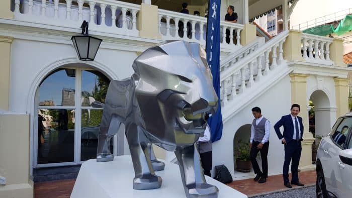 Peugeot lion fiberglass advertising sculpture 3 Metalowy posąg lwa na tle białego budynku z kolumnadą i schodami, przy którym stoją elegancko ubrani ludzie.