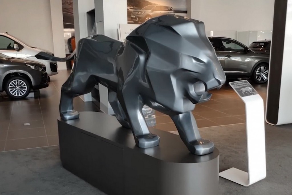 oryg_064490685_1129949550544944_1465385417682452480_o A large, stylized lion sculpture is displayed indoors in a modern car showroom, with several cars visible in the background.