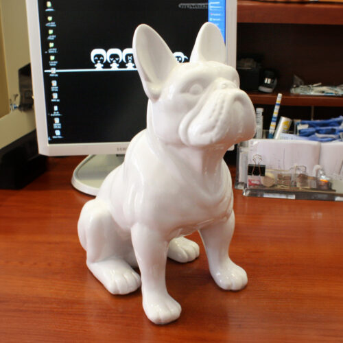 A white ceramic French bulldog statue sits on a wooden desk, with a computer monitor and desk organizer in the background.