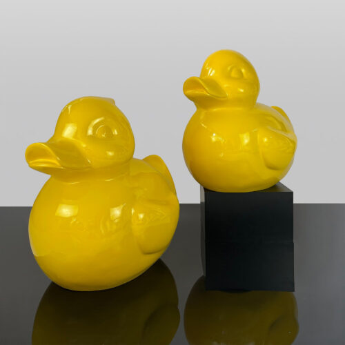 Two yellow duck sculptures, one standing on a black cube and the other directly on a reflective surface.