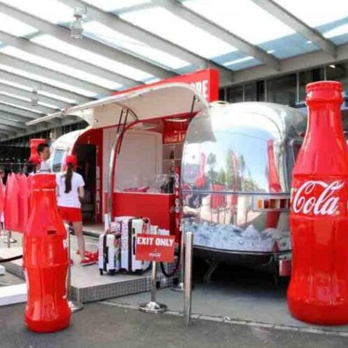 Outdoor Coca-Cola merchandise store with large Coke bottle standees, clothing racks, and a reflective trailer backdrop.