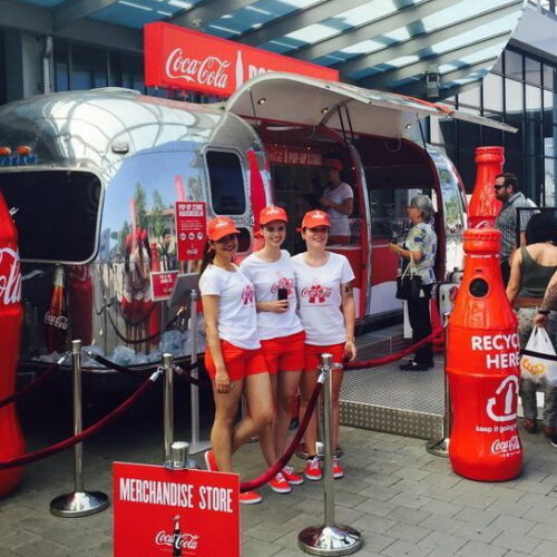 Coca-Cola-themed merchandise store with three women in branded uniforms standing in front of a trailer, surrounded by large Coca-Cola bottle replicas and a recycling station.