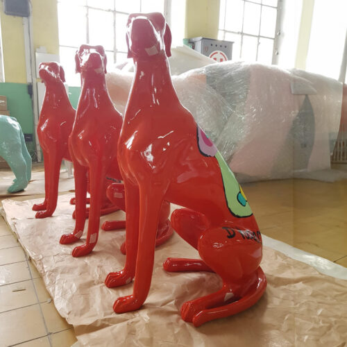 Brightly colored, glossy orange dog sculptures sitting in a row on a paper-covered floor.