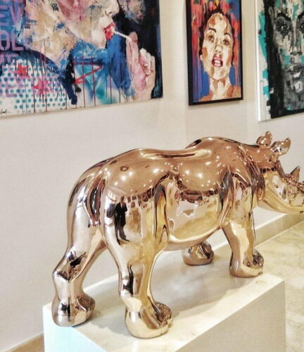 A shiny gold rhinoceros sculpture displayed on a pedestal in an art gallery, with colorful abstract paintings hanging on the wall behind it.